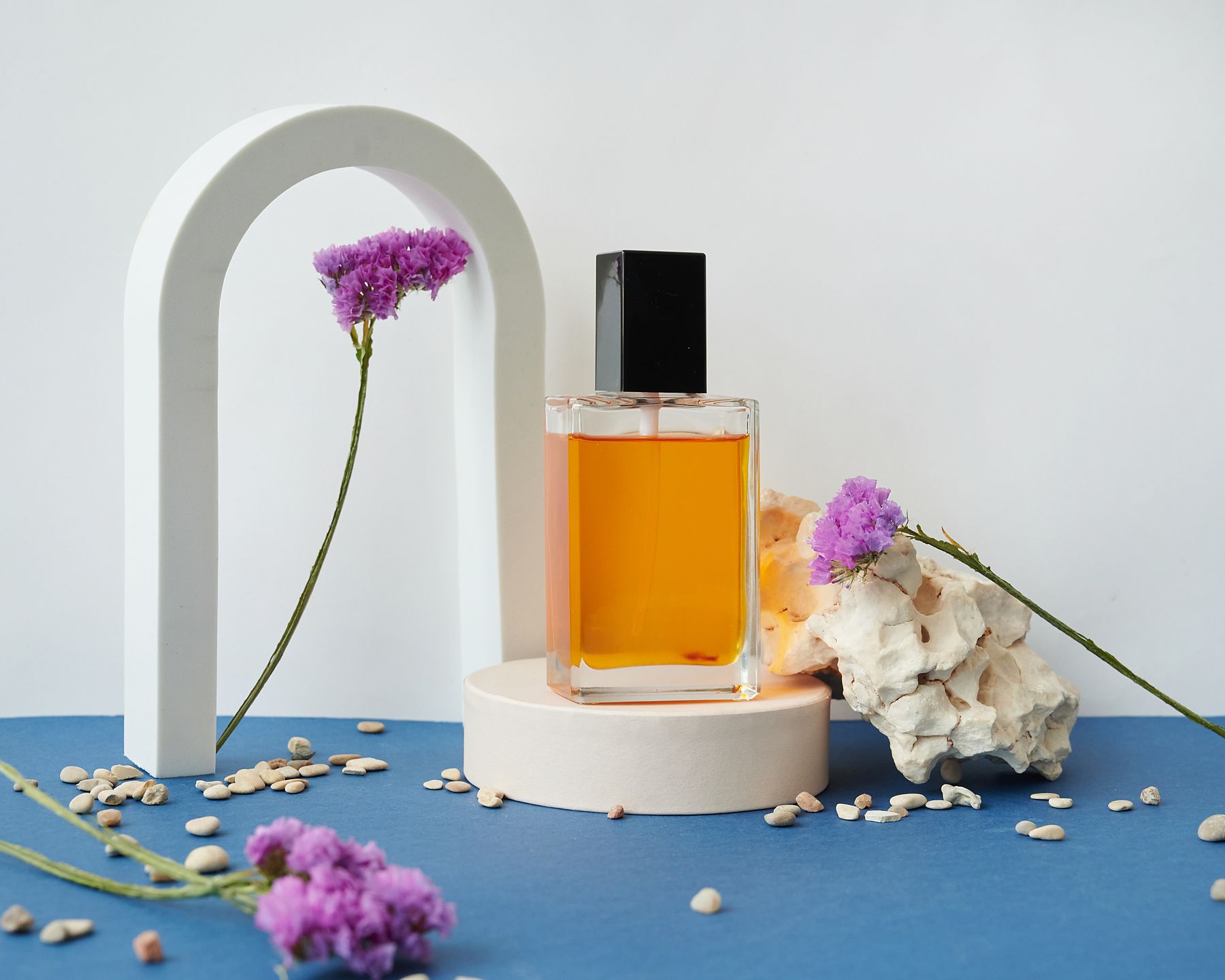 flacon de parfum posé sur un support avec des fleurs mauves séchées, fond bleu et arche blanche
