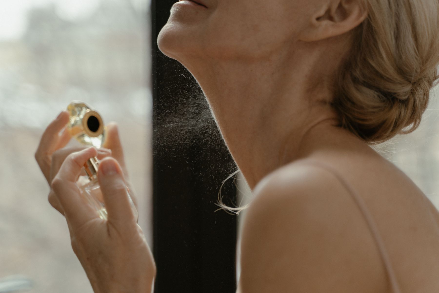 femme qui vaporise du parfum sur son cou près d'une fenêtre, lumière naturelle